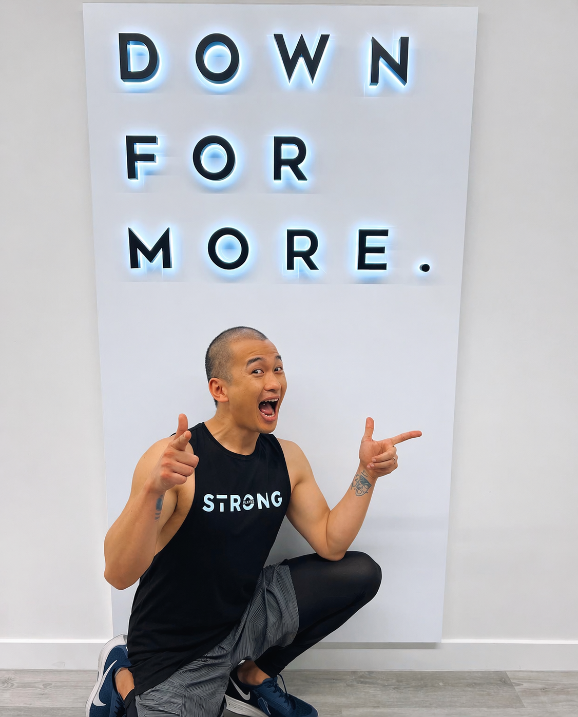 Tim wearing a ‘STRONG’ tank top pointing towards a motivational sign indoors.
